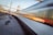 A high-speed train blurs past on a railway track, surrounded by a network of electric cables and infrastructural elements. The motion of the train creates a dynamic visual effect, contrasting with the static elements of the railway environment, such as tracks, overhead wires, and support poles.