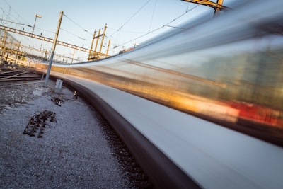 A high-speed train blurs past on a railway track, surrounded by a network of electric cables and infrastructural elements. The motion of the train creates a dynamic visual effect, contrasting with the static elements of the railway environment, such as tracks, overhead wires, and support poles.