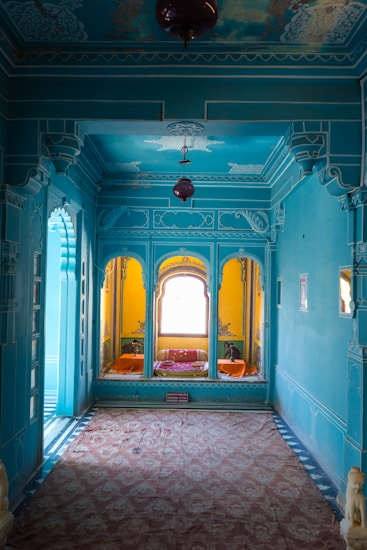 An ornately decorated room features vibrant blue walls with detailed engravings and moldings. A large window at the rear illuminates a cozy seating area adorned with colorful pillows and bright yellow walls. The ceiling also showcases intricate patterns, and a red hanging lamp adds to the decor. The floor is covered with an elaborate carpet, adding a further touch of elegance.
