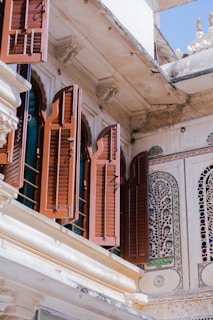 An exterior architectural detail featuring intricately carved wooden shutters on a building. The open shutters reveal arched window frames. The facade is adorned with ornate stone carvings and colorful mosaic patterns.