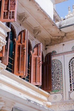 An exterior architectural detail featuring intricately carved wooden shutters on a building. The open shutters reveal arched window frames. The facade is adorned with ornate stone carvings and colorful mosaic patterns.