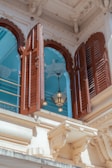 A richly detailed architectural element featuring ornate brown wooden shutters on a white building facade. Behind the shutters, an interior with a light blue ceiling and decorative patterns is visible. A pendant light with a metal frame and glass panels hangs in the opening.