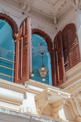 A richly detailed architectural element featuring ornate brown wooden shutters on a white building facade. Behind the shutters, an interior with a light blue ceiling and decorative patterns is visible. A pendant light with a metal frame and glass panels hangs in the opening.