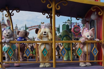 Several costumed characters are standing on a colorful float in what appears to be a theme park with a castle and crowd of people visible in the background. The characters are in various fun and playful poses with vibrant decorations around them.