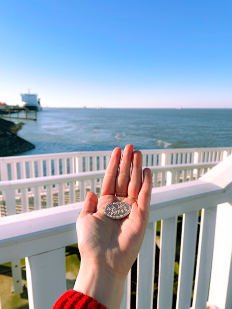 A hand wearing a red sweater holds a coin against the backdrop of a bright, sunny seascape. A white wooden railing is prominently visible, with the sea and sky extending into the distance. A large ship is seen on the left side, near the horizon.