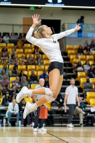A female volleyball player is captured in mid-air, poised to serve the ball during a match. She is wearing a white jersey and black shorts, with knee pads and white sneakers. The background shows an indoor sports arena filled with spectators seated in yellow seats.