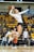 A female volleyball player is captured in mid-air, poised to serve the ball during a match. She is wearing a white jersey and black shorts, with knee pads and white sneakers. The background shows an indoor sports arena filled with spectators seated in yellow seats.
