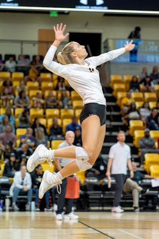 A female volleyball player is captured in mid-air, poised to serve the ball during a match. She is wearing a white jersey and black shorts, with knee pads and white sneakers. The background shows an indoor sports arena filled with spectators seated in yellow seats.