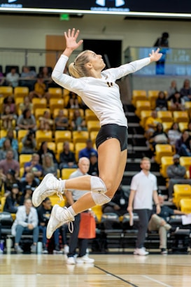 A female volleyball player is captured in mid-air, poised to serve the ball during a match. She is wearing a white jersey and black shorts, with knee pads and white sneakers. The background shows an indoor sports arena filled with spectators seated in yellow seats.