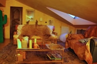 A cozy living room with warm lighting and rustic decor, featuring a large brown armchair, a decorative cactus, various candles on a wooden table, and a fur throw. The room is under a slanted ceiling with a skylight, and includes wooden floors and eclectic decorations including a framed picture and a radiator.