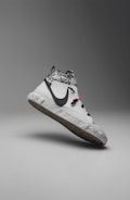 A modern sneaker with a sleek, minimalist design. It features a predominantly white color with a contrasting black swoosh logo on the side. The upper section displays a gray and white camouflage pattern, while the sole has a recycled, speckled texture. There are distinctive black laces and a small red detail near the top.