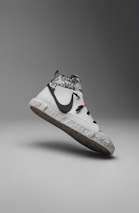 A modern sneaker with a sleek, minimalist design. It features a predominantly white color with a contrasting black swoosh logo on the side. The upper section displays a gray and white camouflage pattern, while the sole has a recycled, speckled texture. There are distinctive black laces and a small red detail near the top.