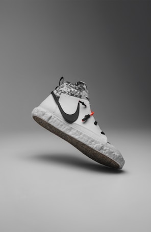 A modern sneaker with a sleek, minimalist design. It features a predominantly white color with a contrasting black swoosh logo on the side. The upper section displays a gray and white camouflage pattern, while the sole has a recycled, speckled texture. There are distinctive black laces and a small red detail near the top.