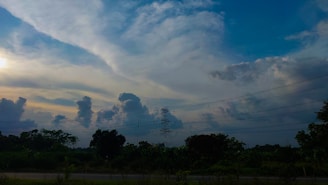A scenic landscape featuring vast clouds in the sky with a gradient of colors from bright blue to darker shades. A transmission tower stands prominently against the sky, surrounded by lush greenery and trees below. Power lines stretch across the scene, adding an element of infrastructure to the natural setting.