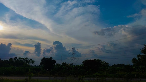 A scenic landscape featuring vast clouds in the sky with a gradient of colors from bright blue to darker shades. A transmission tower stands prominently against the sky, surrounded by lush greenery and trees below. Power lines stretch across the scene, adding an element of infrastructure to the natural setting.