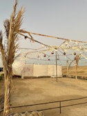 A simple outdoor tent structure with metal frames and fabric covering. The area is surrounded by a sandy landscape with sparse vegetation. The sky is clear and there are rustic elements like hanging ornaments and ropes adorning the frame.