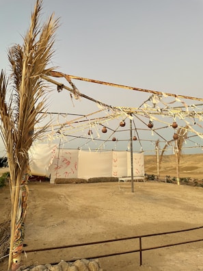 A simple outdoor tent structure with metal frames and fabric covering. The area is surrounded by a sandy landscape with sparse vegetation. The sky is clear and there are rustic elements like hanging ornaments and ropes adorning the frame.