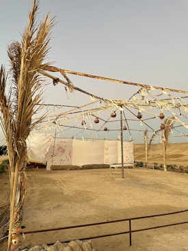 A simple outdoor tent structure with metal frames and fabric covering. The area is surrounded by a sandy landscape with sparse vegetation. The sky is clear and there are rustic elements like hanging ornaments and ropes adorning the frame.