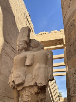 An ancient, weathered stone statue of a figure with a tall headdress stands amidst temple ruins. The surrounding walls and columns are covered in hieroglyphic carvings, and the blue sky adds contrast above.