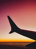 A sleek airplane wing cutting through a vibrant sunset sky with hues of orange and purple.