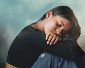 A woman is sitting with her head resting on her arms, eyes closed, and appears to be in deep thought or possibly feeling sad. She is wearing a black long-sleeve shirt and blue jeans with a small tear. The background is a light, cloudy sky, giving a serene yet melancholic atmosphere.