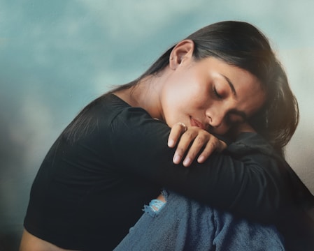 A woman is sitting with her head resting on her arms, eyes closed, and appears to be in deep thought or possibly feeling sad. She is wearing a black long-sleeve shirt and blue jeans with a small tear. The background is a light, cloudy sky, giving a serene yet melancholic atmosphere.