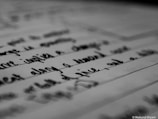 A close-up view of handwritten text on a piece of paper, captured in black and white. The emphasis is on the script, with some areas blurred, likely due to depth of field.
