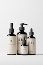 Four minimalist cosmetic bottles with black caps and beige labels displaying the brand 'LBL'. The bottles vary in size and design, featuring pump and dropper caps. They are placed against a clean, white background.
