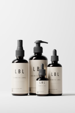 Four minimalist cosmetic bottles with black caps and beige labels displaying the brand 'LBL'. The bottles vary in size and design, featuring pump and dropper caps. They are placed against a clean, white background.