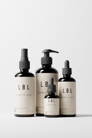 Four minimalist cosmetic bottles with black caps and beige labels displaying the brand 'LBL'. The bottles vary in size and design, featuring pump and dropper caps. They are placed against a clean, white background.