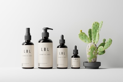 A minimalist arrangement of cosmetic bottles labeled 'LBL' is positioned in a line on a surface. Next to them sits a small potted cactus plant with green pads and hints of brown spots. The background is a light, neutral color adding to the clean aesthetic.