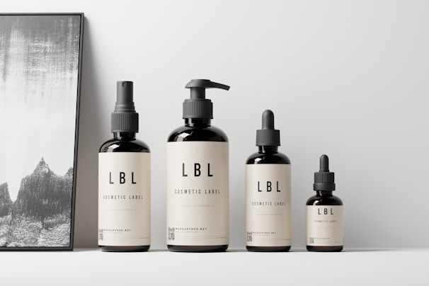 A set of four cosmetic bottles with minimalist labels displayed next to a framed abstract artwork. The bottles vary in size and feature pumps and droppers. The setting is clean and modern, with a neutral color palette.