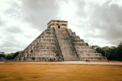 A large, ancient stone pyramid with stepped sides stands prominently under a cloudy sky. The pyramid is surrounded by open grassy areas and a few trees. Several people are gathered at the base, showcasing the structure's massive scale and historical significance.