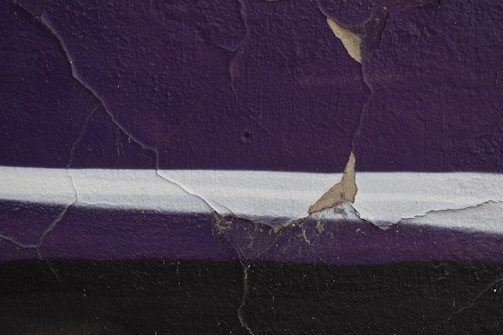 A close-up of a wall with significant cracks in the purple paint. The wall features a white horizontal stripe in the middle. There are patches where the paint is peeling off, revealing a beige color underneath.