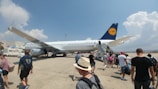 A group of people boarding a large commercial airplane on the tarmac. The airplane, with a visible Lufthansa logo on its tail, is parked under a bright blue sky with some scattered clouds. Several individuals are ascending the boarding stairs, while others are approaching the aircraft from the tarmac, carrying luggage and wearing casual summer clothing.