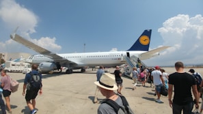 A group of people boarding a large commercial airplane on the tarmac. The airplane, with a visible Lufthansa logo on its tail, is parked under a bright blue sky with some scattered clouds. Several individuals are ascending the boarding stairs, while others are approaching the aircraft from the tarmac, carrying luggage and wearing casual summer clothing.