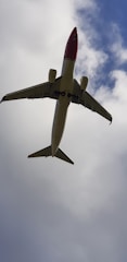 A passenger airplane is seen from below, flying against a backdrop of blue sky partially covered with clouds. The plane has a white body with a red nose and is positioned in the center of the image.