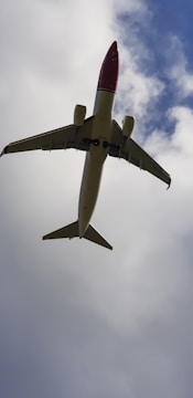 A passenger airplane is seen from below, flying against a backdrop of blue sky partially covered with clouds. The plane has a white body with a red nose and is positioned in the center of the image.