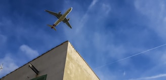 An airplane is flying high in the sky above a corner of a building. The building is beige with a green window shutter, and there's a security camera mounted on the wall. The sky is clear, with a few wispy clouds.