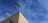 An airplane is flying high in the sky above a corner of a building. The building is beige with a green window shutter, and there's a security camera mounted on the wall. The sky is clear, with a few wispy clouds.