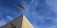 An airplane is flying high in the sky above a corner of a building. The building is beige with a green window shutter, and there's a security camera mounted on the wall. The sky is clear, with a few wispy clouds.