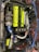 A detailed view of a car engine with a prominent neon yellow cover labeled 'Twin Cam 16 Super Charger' and 'Toyota'. Surrounding this are various components, including a turbocharger, hoses, and wires. There are several branded parts, such as a blue intake pipe with 'Ultra Racing' printed on it.
