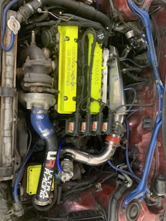 A detailed view of a car engine with a prominent neon yellow cover labeled 'Twin Cam 16 Super Charger' and 'Toyota'. Surrounding this are various components, including a turbocharger, hoses, and wires. There are several branded parts, such as a blue intake pipe with 'Ultra Racing' printed on it.