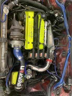A detailed view of a car engine with a prominent neon yellow cover labeled 'Twin Cam 16 Super Charger' and 'Toyota'. Surrounding this are various components, including a turbocharger, hoses, and wires. There are several branded parts, such as a blue intake pipe with 'Ultra Racing' printed on it.