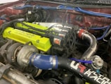 A detailed view of a car engine setup showcasing a turbocharger system. The engine features a bright lime-green valve cover with visible branding, blue silicone hoses marked with logos, and polished metal piping. Several ignition coils and sleek wiring are also present, indicating a performance-oriented configuration.