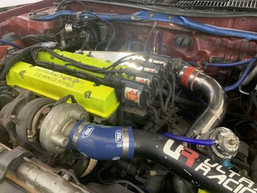 A detailed view of a car engine setup showcasing a turbocharger system. The engine features a bright lime-green valve cover with visible branding, blue silicone hoses marked with logos, and polished metal piping. Several ignition coils and sleek wiring are also present, indicating a performance-oriented configuration.
