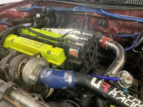 A detailed view of a car engine setup showcasing a turbocharger system. The engine features a bright lime-green valve cover with visible branding, blue silicone hoses marked with logos, and polished metal piping. Several ignition coils and sleek wiring are also present, indicating a performance-oriented configuration.