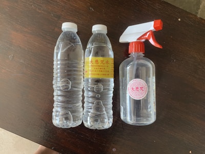 On a dark wooden surface, there are two clear plastic bottles with white caps, and a transparent spray bottle with a red and white nozzle. The plastic bottles have yellow labels with red and black text. The spray bottle has a red cap with a white label bearing circular red designs.