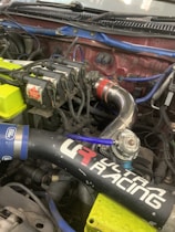 A close-up view of a car engine with visible components including ignition coils marked with 'Bosch Coil', various pipes, and wires. There is a prominent black air intake tube with 'Ultra Racing' branding and colorful wires in blue and yellow. A metallic pipe featuring a red connector is also visible.
