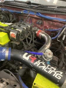 A close-up view of a car engine with visible components including ignition coils marked with 'Bosch Coil', various pipes, and wires. There is a prominent black air intake tube with 'Ultra Racing' branding and colorful wires in blue and yellow. A metallic pipe featuring a red connector is also visible.
